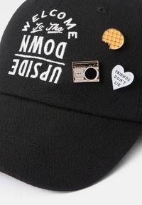 Melna cepure ar baltiem izšūtiem vārdiem "LAIPNI NĀC UN PAGRIEZIES PA PUSI". Cepurei ir trīs piespraudes: retro kamera, koduma vafeļu gabals un sirds ar tekstu.