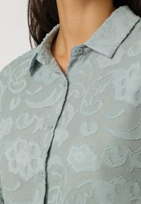 Blusa azul claro con un patrón texturizado de bordado floral y pequeños botones. Material suave y diseño de cuello tipo camisa.