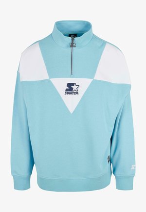 Hele sinine fliistepusa, millel on pooltagasitõmbamine, valged kolmnurksed aktsendid ja must logo rindkere piirkonnas. Pehme tekstuur ja lõtv istuvus.