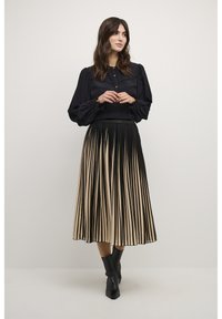 Schwarze Bluse mit Puffärmeln und Knöpfen, kombiniert mit einem plissierten Rock mit schwarz-beigen vertikalen Streifen. Schwarze Knöchelstiefel.