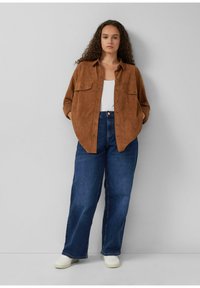 Frau mit lockigem Haar, die ein braunes Hemd mit Knöpfen über einem weißen Oberteil, blaue Jeans und weiße Sneakers trägt, vor einem schlichten grauen Hintergrund steht.