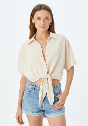 Chemise cintrée à manches courtes beige clair avec un design à boutons et un nœud sur le devant, associée à un short en denim déchiré. Tissu doux, coupe décontractée.