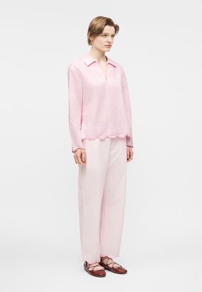 Nő áll, halvány rózsaszín hosszú ujjú blúzt és hozzáillő, széles szárú nadrágot visel hullámos szegélyekkel, sötétvörös, fűzős lapos cipővel párosítva.