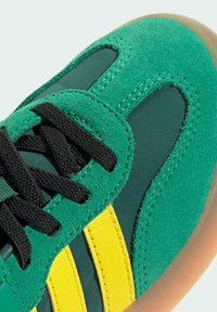 Gröna mockasneakers med svarta snören, gula läderremsor, mörkgröna tygpaneler och en sula av gummigummi, närbild.