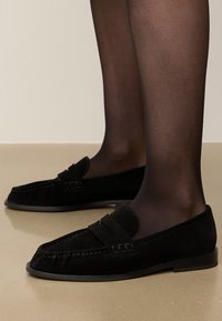 Zwarte nubuck instappers met een ronde neus, decoratieve band en stikdetails, gecombineerd met doorzichtige zwarte panty, gepositioneerd op een reflecterend oppervlak.