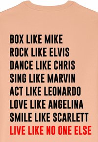 T-shirt pêche avec un texte noir et rouge audacieux comportant des phrases motivantes empilées verticalement au dos. Tissu en coton doux, design à col rond.