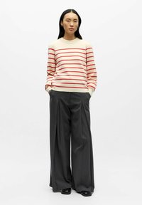 Maglione color crema con strisce rosse, abbinato a pantaloni grigi a gamba larga, con una texture liscia e una vestibilità rilassata.