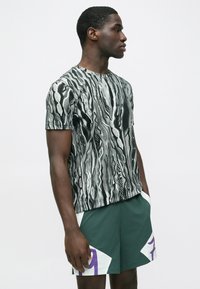 Męski t-shirt z krótkim rękawem w czarno-biały wzór z abstrakcyjnymi kształtami; noszony z zielonymi szortami z białymi i fioletowymi akcentami.