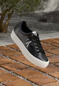 Zapatilla negra de cuero con suela de goma blanca, que cuenta con cordones redondos y detalle de logo en el lateral y en la lengüeta. Patrón texturizado en la superficie.