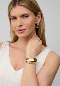 Złote kolczyki w kształcie kół oraz masywna złota bransoleta noszone przez modelkę z długimi blond włosami, na neutralnym tle.