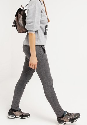 Un pantalon de survêtement gris associé à un sweat-shirt gris clair arborant un logo, des chaussures de sport noires avec un motif camouflage, et un sac à dos sombre.