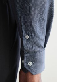 Camisa azul de manga larga con un micropatrón texturizado, con botones blancos y puños ajustados. La tela parece ligera y suave.