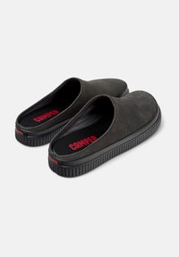 Zapatos sin cordones de ante negro con una suela de goma texturizada, que presentan un logo rojo de "Camper" en la plantilla y el talón. Diseño redondeado y minimalista.