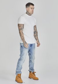 Tricou alb cu dungi, blugi albaştri decolorați și botine bej cu şireturi. Tatuaje vizibile pe brațe, poziție în picioare cu un fundal neutru.