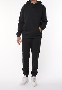 Svart huvtröja och matchande joggers, tillverkade av mjukt material, med en framficka, ribbade ärmslut och vita sneakers.