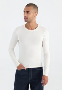 Hosszúujjú kerek nyakú ing, természetes fehér színben, könnyű anyagból készült, sima textúrával. Sötét farmernadrággal párosítva.