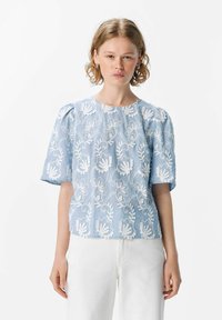 Blusa bordada de color azul claro con patrones florales y mangas cortas. Está hecha de una tela texturizada, con escote redondo y corte relajado.