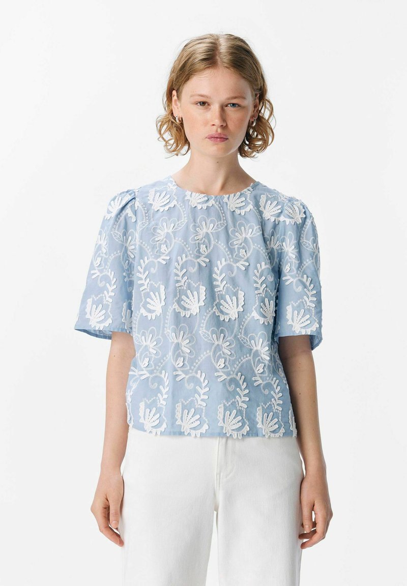 Blusa bordada de color azul claro con patrones florales y mangas cortas. Está hecha de una tela texturizada, con escote redondo y corte relajado.