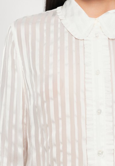 Biała i jasnobeżowa bluzka w paski z kołnierzykiem, z przodu zapinana na guziki, z falbaniastymi detalami wzdłuż plisy. Miękka, przezroczysta tkanina.