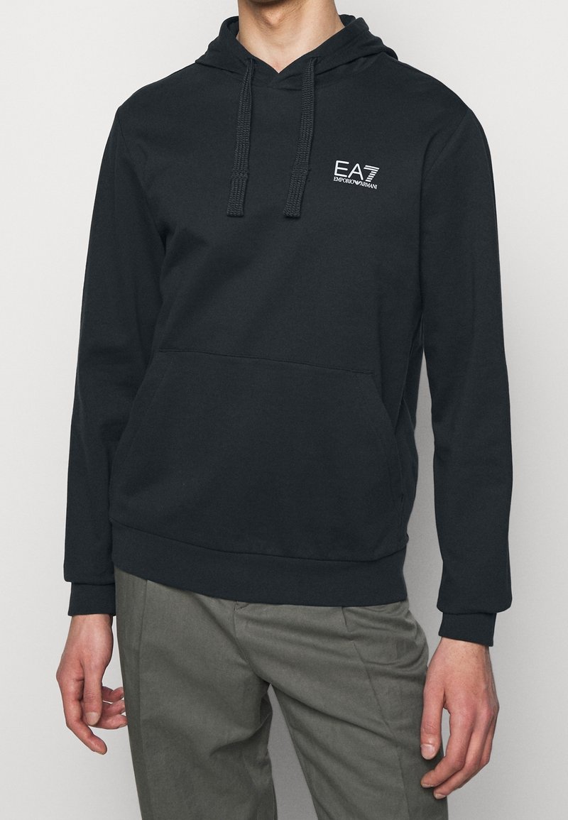 Mies, joka seisoo rento kädet sivuilla, päällä musta huppari, jossa on EA7 Emporio Armani -logo rinnassa ja harmaat housut.