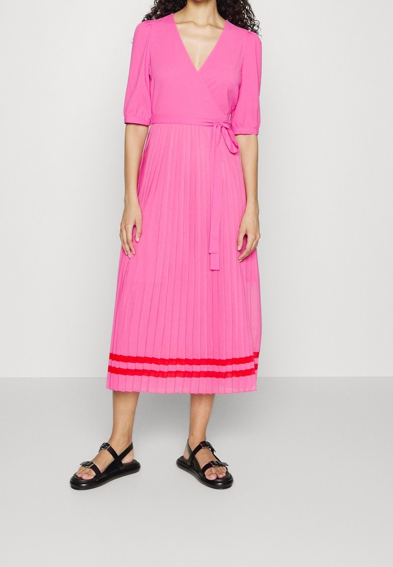 Robe midi plissée rose avec un décolleté en V et des manches courtes, dotée d'un lien à la taille et de rayures rouges sur l'ourlet. Portée avec des sandales noires.