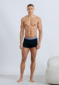 Φορώντας μόνο μαύρο εσώρουχο τύπου μποξέρ με γκρι ζώνη μέσης, ένας καλοντυμένος νεαρός άνδρας στέκεται ξυπόλητος μπροστά σε απλό φόντο.