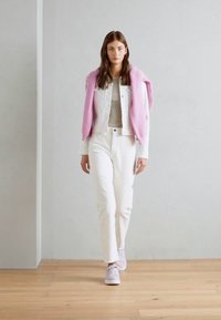 Giacca di denim bianca sopra una maglietta grigia chiara, abbinata a pantaloni bianchi a vita alta e un maglione rosa appoggiato sulle spalle. Sneakers rosa chiaro.