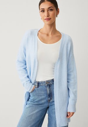 Frau trägt eine hellblaue, offene Strickjacke, ein weißes Top mit Rundhalsausschnitt und blaue Jeans mit hoher Taille, Hände in den Taschen, neutraler Hintergrund.