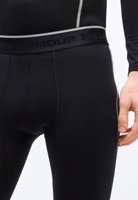 Κοντινό πλάνο μαύρων κολάν συμπίεσης Under Armour με ευδιάκριτες ραφές και λογότυπο στη μέση, σε μερικώς εμφανιζόμενο άτομο.