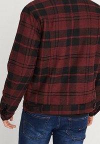 Rød og sort ternet flanneljakke med en blød tekstur. Har knapdetaljer og et lige snit, båret med blå jeans.