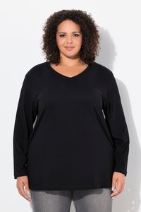 Femme aux cheveux bouclés portant un haut noir à manches longues avec un col en V et un jean gris, debout sur un fond blanc, faisant face à l'avant.