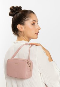 Borsa a tracolla in pelle rosa con una texture liscia, forma rettangolare, chiusura superiore a zip e tracolla sottile. Foulard dorato sul davanti.