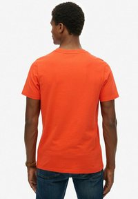 Pomarańczowa bawełniana koszulka z okrągłym dekoltem i krótkimi rękawami, widok z tyłu. Gładka struktura, prosty fason i brak widocznych wzorów.