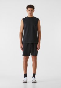 Schwarzes ärmelloses Top kombiniert mit schwarzen Shorts. Der Stoff wirkt glatt, mit einer lässigen Passform und einem Kordelzugbund. Die Schuhe haben ein Design in Weiß und Grau.