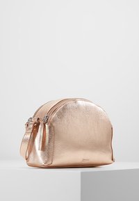 Torebka w kolorze różowego złota o zaokrąglonym kształcie, z dwiema komorami na zamek błyskawiczny i cienkim paskiem na ramię. Teksturalna powierzchnia dodaje wymiaru.