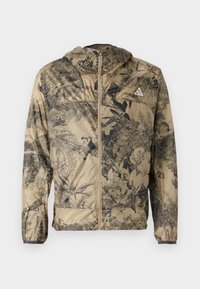 Leichte beigefarbene Jacke mit schwarzem botanischen Druck. Verfügt über eine Kapuze, einen Frontreißverschluss und elastische Bündchen. Minimalistisches Design ohne sichtbare Hardware.