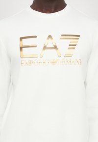 Sweatshirt branco com um logótipo impresso a dourado com "EA7 EMPORIO ARMANI." Textura suave e decote redondo. Mangas longas sem detalhes adicionais.