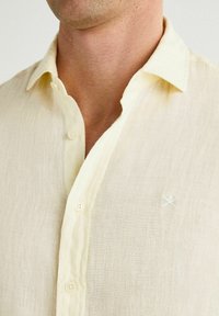 Camisa de lino color beige claro con cuello, que presenta una fila de botones y un sutil bordado en el pecho.