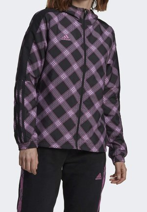 Person trägt eine schwarz-rosa karierte Adidas Zip-Up-Jacke und schwarze Hose mit pinkfarbenen Adidas-Logos.