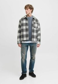 Mand iført grå og hvid ternet jakke, blå sweatshirt, falmede blå jeans, sorte sko og sølvkæde, stående og kigger til venstre.