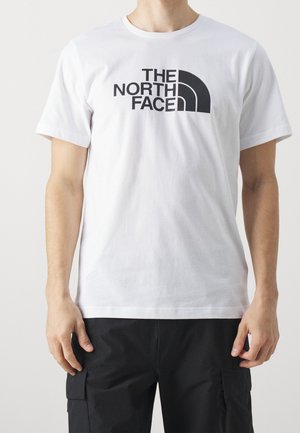 Mężczyzna w białej koszulce z krótkim rękawem z czarnym logo "THE NORTH FACE" i czarnych spodniach typu cargo, stojący na jednolitym tle.