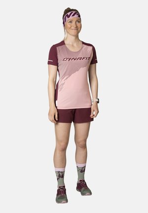 Vrouw staat gekleed in roze en bordeaux Dynafit sportkleding, patroon hiking sokken, trail schoenen en een bijpassende hoofdband.