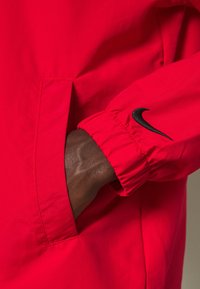 Rode lichtgewicht jas met elastische manchetten, voorzien van een zijzak en een zwart Nike swoosh logo op de mouw. Gladde stoftextuur.