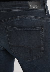 Mörkblå denimjeans har en bakficka med en sömdesign i kurvor. Midjebandet har en svart läderetikett med texten "Mavi Jeans Co."