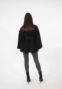 Sort kåbestil-kappe med en snørret talje, båret over grå skinny jeans. Modellen står med ryggen mod, har langt, glat hår og sorte hæle.