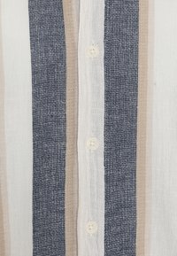 Koszula lniana z pionowymi paskami w kolorach granatowym, beżowym i kremowym. Cztery guziki biegnące wzdłuż przodu, teksturowana tkanina o wplecionym wzorze.