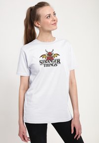 Hvid bomuld T-shirt med et grafisk design af et rødt væsen med horn og vinger samt teksten "STRANGER THINGS" i fede sorte bogstaver.