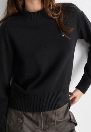 Osoba ubrana w czarny prążkowany sweter z okrągłym dekoltem i brązowe spodnie z kieszeniami na klapki zapinanymi na guziki, stojąca z jedną ręką na biodrze.