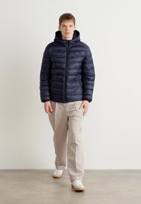 Giubbotto imbottito blu navy con zip frontale e cappuccio. Presenta un motivo a righe orizzontali e un tessuto liscio. Abbinato a pantaloni beige e scarpe bianche.