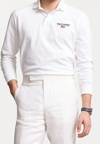 Vit långärmad pikétröja med knäppt krage och blå/röd brodering av "POLO SPORT". Kombinerad med ljusa byxor och en klocka.